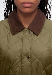 Gros plan d'une personne portant une veste matelassée vert olive avec un col en velours côtelé marron et des boutons-pression argentés.