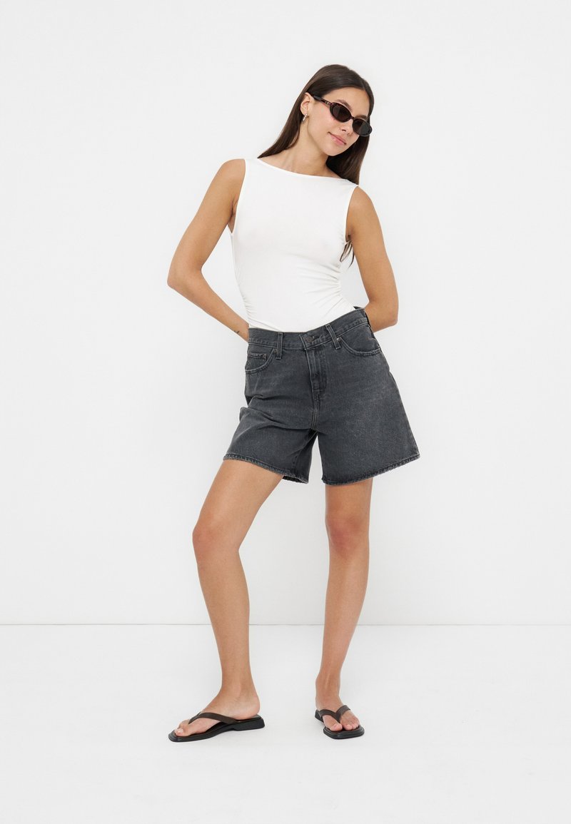 Femme portant un débardeur blanc sans manches, un short en jean noir, des tongs noires et des lunettes de soleil, posant devant un fond blanc.
