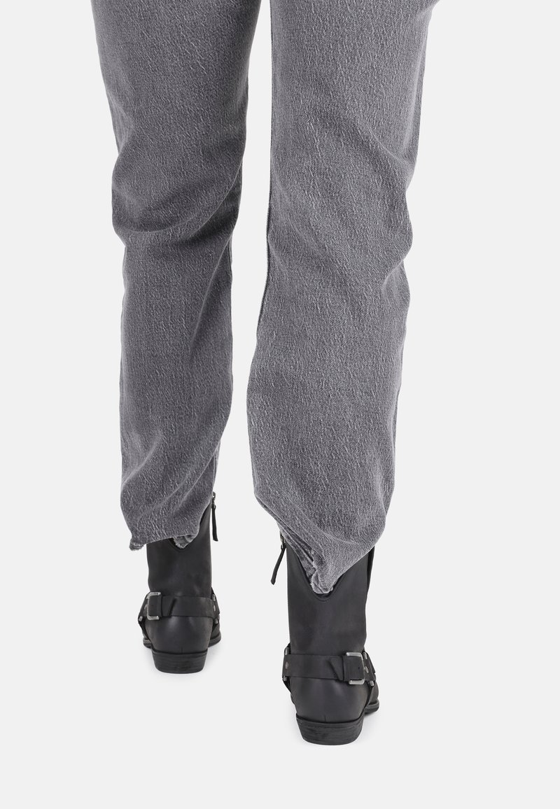 Osoba ubrana w szare, teksturowane spodnie wsadzone w czarne botki za kostkę z klamrami, stojąca na tle białym.