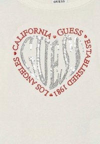 Maglione off-white con un design a forma di cuore in paillettes con la scritta "GUESS" e "CALIFORNIA LOS ANGELES ESTABLISHED 1981" in testo rosso.