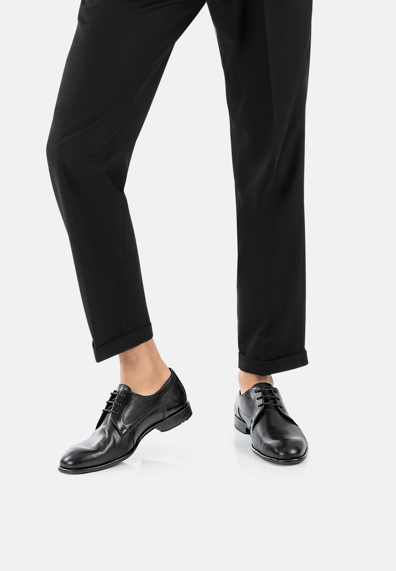 Schwarze Anzugschuhe aus strukturiertem Leder, mit runden Zehen und Schnürsenkeln. Die Hose ist schwarz, an den Knöcheln umgeschlagen und zeigt die Schuhe.
