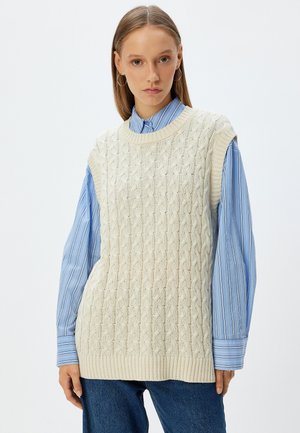 Ivory gebreide mouwloze trui met een kabelpatroon en geribbelde zoom, gelaagd over een blauw gestreept overhemd met knoopsluiting.