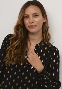 Črna bluzica z zlatimi, ovalnimi pikami, ki ima sproščen ovratnik in naborke na manšetah. Gladek material z rahlim sijajem.