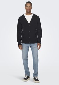 Black knitted cardigan with a V-neck, button closure, ribbed texture, worn over a white shirt, paired with light blue jeans and black sneakers.