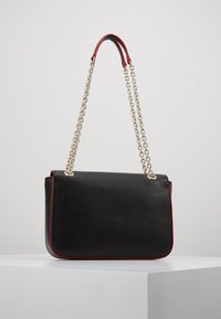 Černá kožená kabelka s červeným lemováním, řetízkovým popruhem na rameno a hladkým povrchem. Obdélníkový tvar a minimalistický design.