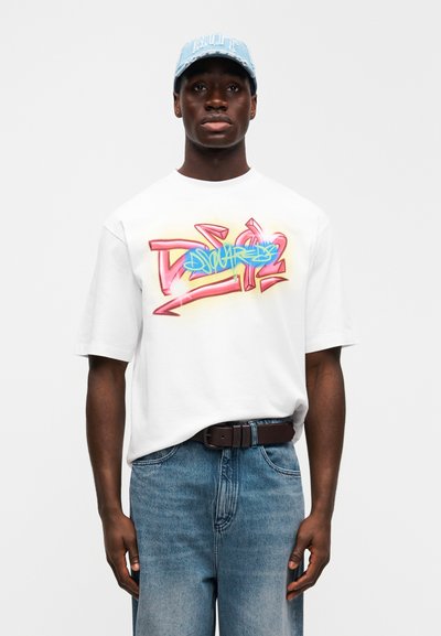 Λευκό graphic t-shirt με πολύχρωμο σχέδιο σε στυλ γκράφιτι, συνδυασμένο με ανοιχτό μπλε τζιν παντελόνι και φθαρμένο μπλε καπέλο. Ορατή καφέ ζώνη.