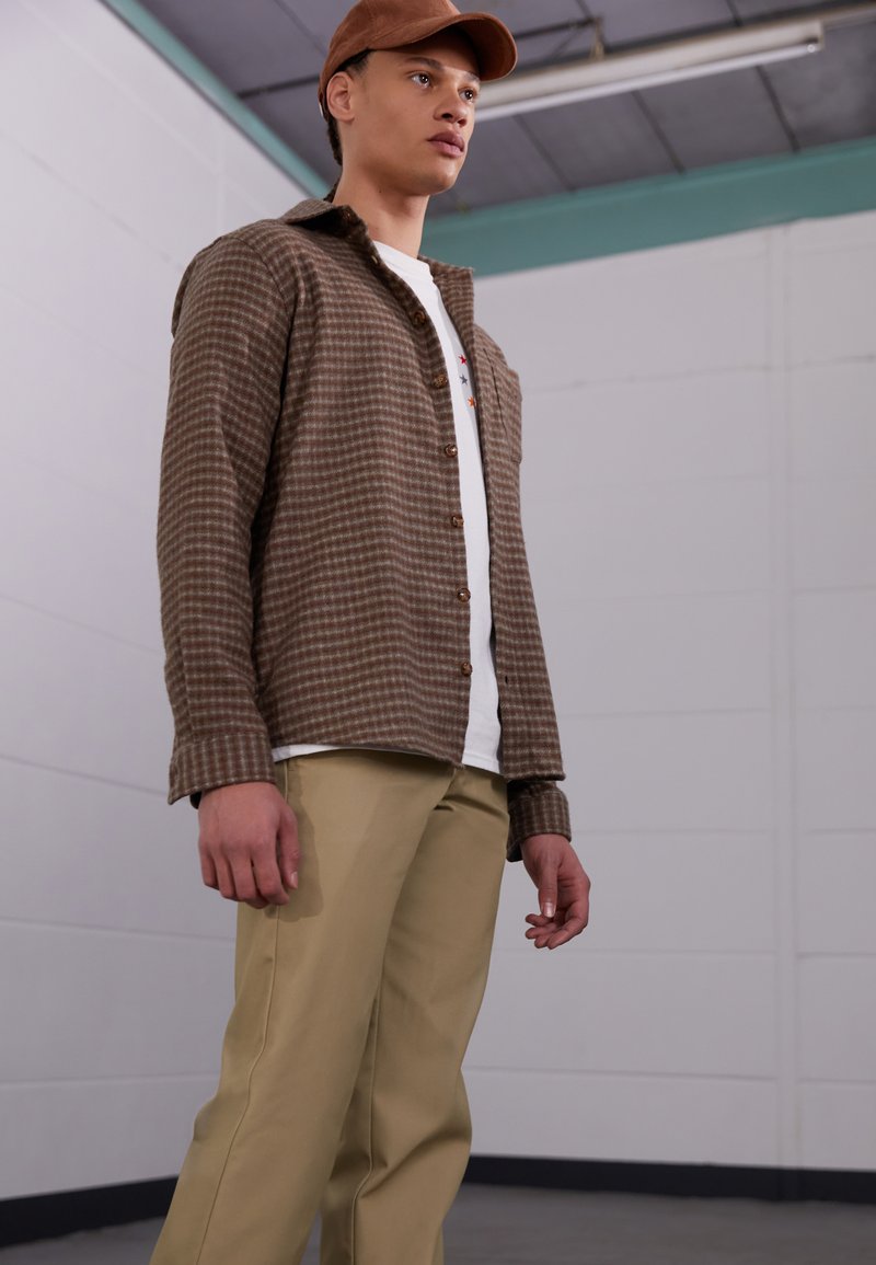 Braunes karierter Hemd mit langen Ärmeln, kombiniert mit beigen Hosen, getragen über einem weißen T-Shirt. Das Modell steht in einer einfachen Inneneinstellung.