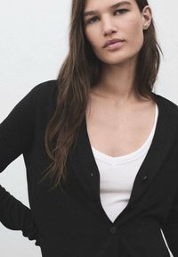 Black cardigan with a v-neck, featuring two buttons and long sleeves, worn over a white top. Soft texture against a neutral background.