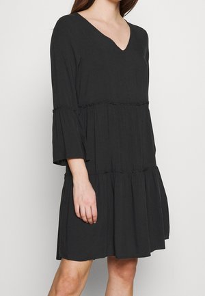 Femme portant une robe noire ample à manches longues et encolure en V, avec une jupe à volants étagée aux détails subtils.