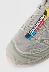 Sapatilha de corrida cinza-claro com parte superior em malha, atacadores elásticos e sola texturizada. Inclui detalhes em vermelho, azul e amarelo na etiqueta da marca.