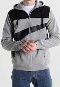 Osoba oblečená v šedé a černé zapínací mikině s diagonálními černými pruhy, ruce složené, a modré džíny viditelné od pasu dolů.