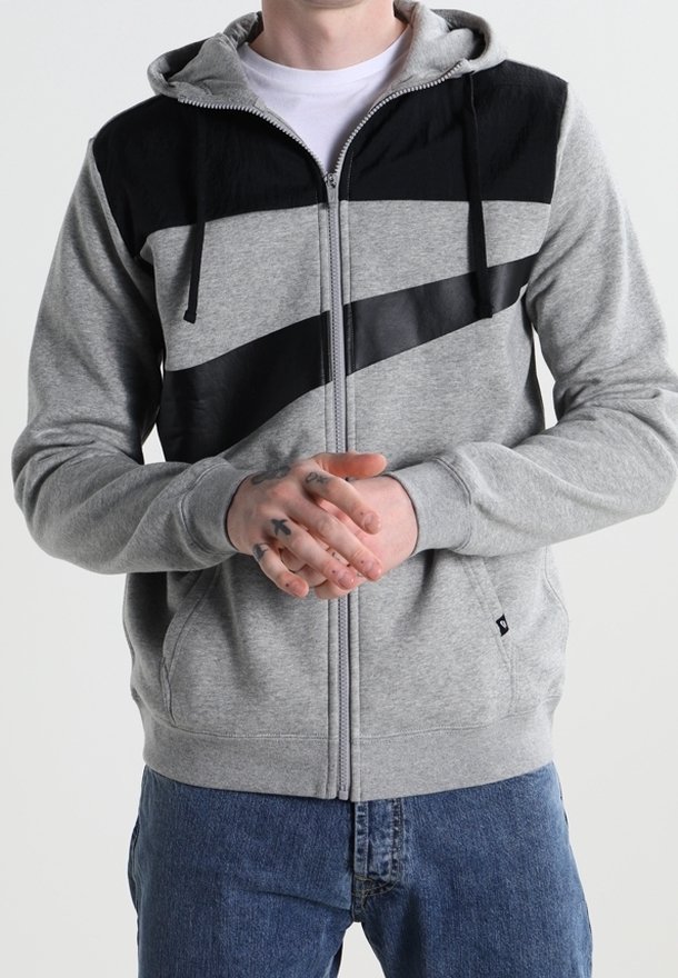 Osoba oblečená v šedé a černé zapínací mikině s diagonálními černými pruhy, ruce složené, a modré džíny viditelné od pasu dolů.