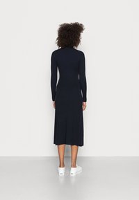 Sovitettu tummansininen ribbimekko, jossa on pitkät hihat ja keski-vartalon pituus, pyöreä kaula-aukko ja hienovarainen leveneminen helmassa. Yhdistetty valkoisiin kenkiin.