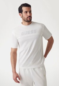 White short-sleeve t-shirt with mesh panel and "GUESS" logo across the chest. Soft fabric, relaxed fit, crew neck design.