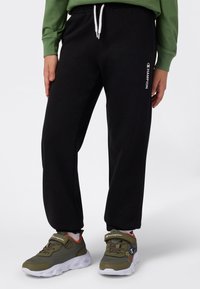 Sorte joggebukser med hvit snøring og side-logo, kombinert med olivengrønne sneakers med hvite såler og borrelås.