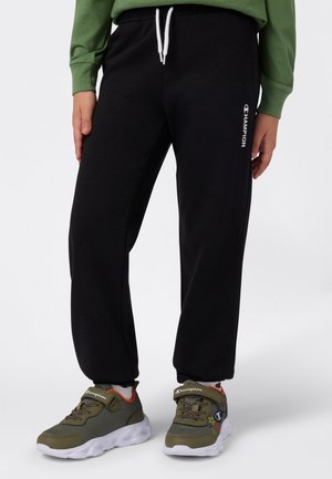 Pantalon de survêtement noir avec cordon blanc et logo sur le côté, associé à des baskets olive avec semelles blanches et bride Velcro.