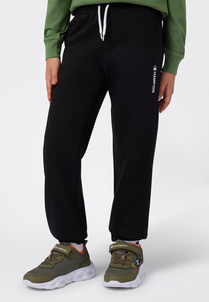 Sorte joggebukser med hvit snøring og side-logo, kombinert med olivengrønne sneakers med hvite såler og borrelås.