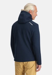 Marineblauwe fleece jas met capuchon, voorzien van een textuurpatroon, ritssluitingen aan de zijkanten, subtiele branding op de schouder en een getailleerde pasvorm.