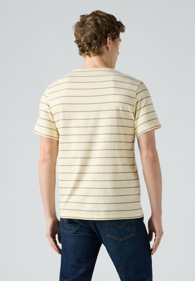 Homme aux cheveux bouclés portant un t-shirt à manches courtes jaune clair avec de fines rayures horizontales sombres et un jean bleu foncé, debout de dos.