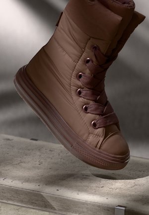 Brązowy sneakers za kostkę z grubą podeszwą i wyściełanym materiałowym wierzchem, pokazany w powietrzu nad betonową powierzchnią.