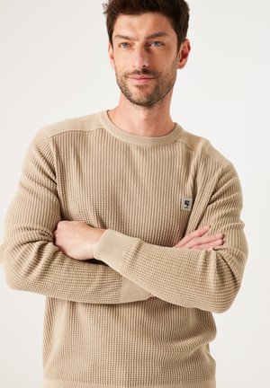 Suéter de punto beige, con un diseño texturizado de canalé, cuello redondo y detalles contrastantes en las mangas, adicionado con un pequeño parche de logo en el pecho.