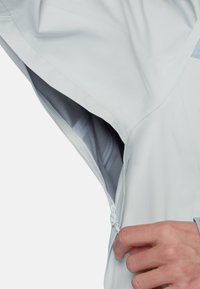 Hand öffnet den Reißverschluss am Ärmel einer grauen Jacke und zeigt das Innenfutter und die Reißverschlussdetails auf dem hellen Außenmaterial.