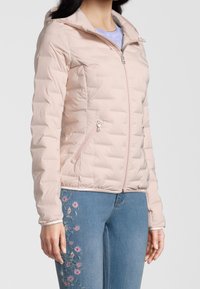 Doudoune rose clair avec fermeture éclair à l'avant, capuche et motif matelassé. Dispose de deux poches latérales zippées. Portée sur un denim avec broderie florale.