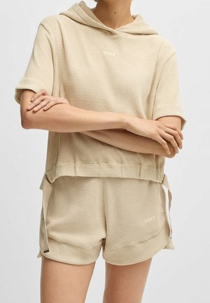 Osoba w beżowej, teksturowanej, krótkim rękawem z kapturem i pasujących do tego krótkich spodenkach z wiązaniami po bokach, z ramionami skrzyżowanymi, twarz niewidoczna.