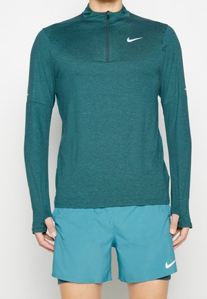 Person trägt ein türkisfarbenes langärmliges Quarter-Zip-Sportoberteil und hellblaue Sportshorts mit weißen Nike-Logos auf beiden Kleidungsstücken.