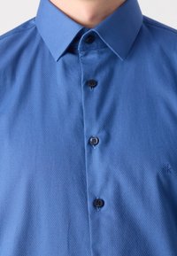 Camicia blu a bottoni con tessuto testurizzato, caratterizzata da un colletto classico, cinque bottoni neri e un piccolo logo sul lato destro in basso.
