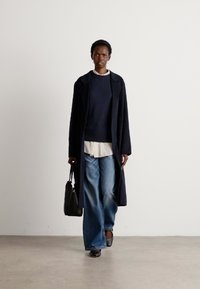 Casaco longo azul marinho sobre um suéter azul marinho e uma camisa branca, combinado com jeans de perna larga e sapatos pretos. Uma bolsa preta completa o visual.