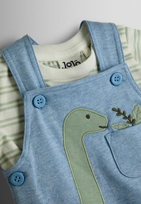 Salopetă albastră pentru bebeluși cu aplică verde în formă de dinozaur și frunze brodate, purtată peste un tricou cu mânecă lungă în dungi albe și verzi.