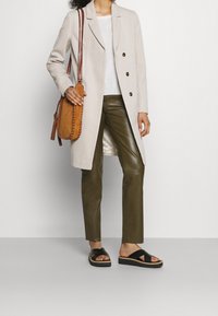 Donna che indossa un lungo cappotto beige, una maglietta bianca, pantaloni in pelle verde oliva, sandali con plateau neri e porta una borsa a tracolla in pelle cucita color tan.