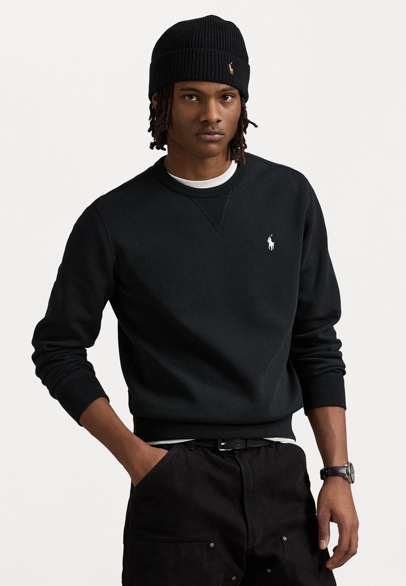 Sort bomulds sweatshirt med ribbede manchetter, der har et lille hvidt logo på venstre bryst. Båret med sorte bukser og en sort ribbede beanie.