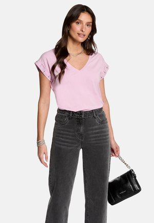 Roze V-hals t-shirt met opgerolde mouwen, versierd met studs, gecombineerd met hooggerimde donkergrijze jeans en een zwarte handtas met een kettinghengsel.