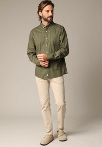 Olijfgroen overhemd met lange mouwen en een enkele borstzak, gecombineerd met crème-kleurige slim-fit pants en beige instappers.