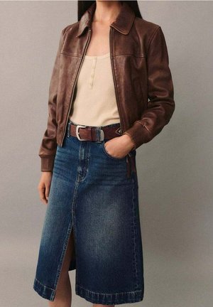 Kobieta w brązowej skórzanej kurtce, beżowej bluzce henley, brązowym pasku i dżinsowej spódnicy sięgającej kolan z przednim rozcięciem, stojąca na jednolitym tle.