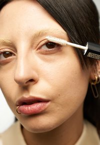 Brosse de mascara transparent appliquée sur les cils, avec une brosse blanche texturée. Le modèle a des sourcils blonds clairs et un anneau de septum. Texture de peau lisse.