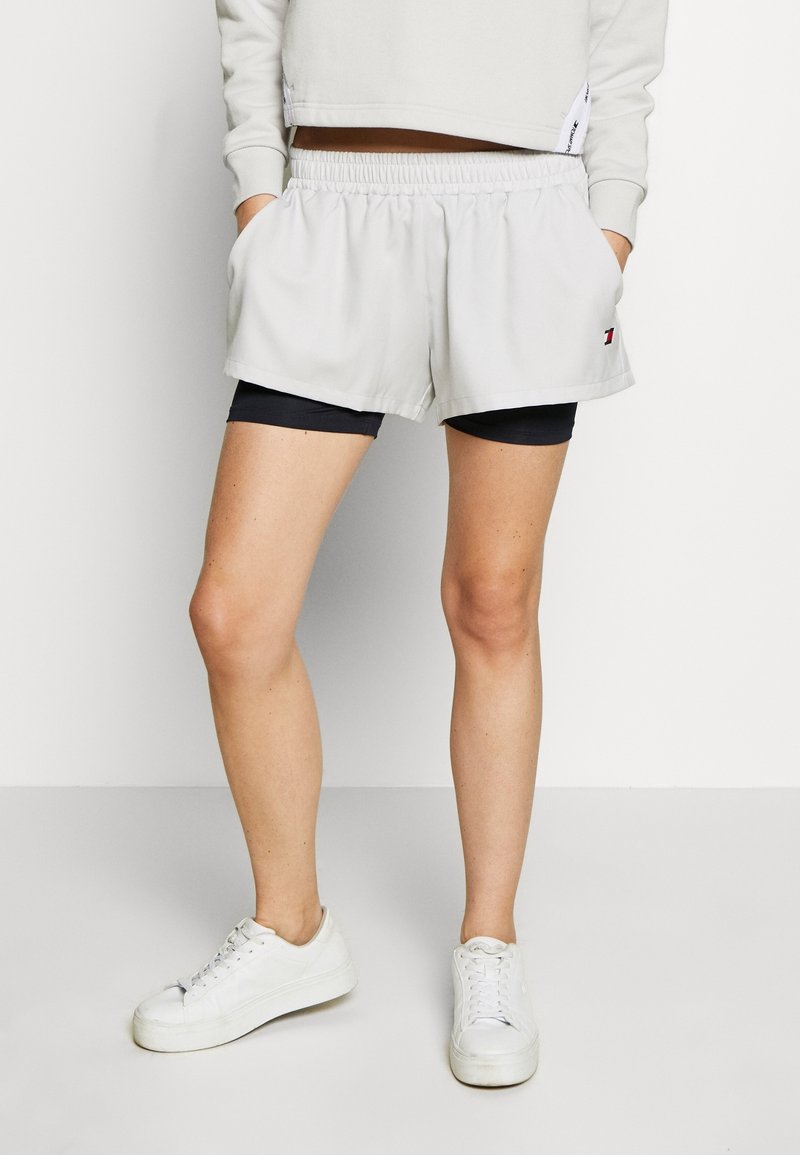 Pantalones cortos deportivos de color gris claro con cintura elástica, bolsillos laterales y un detalle de logotipo. Llevados sobre pantalones cortos de compresión azul marino.