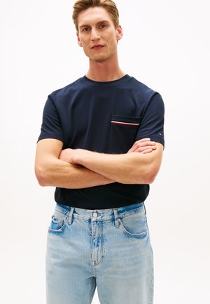 Bărbat cu piele deschisă la culoare și păr blond, purtând un tricou bleumarin cu un detaliu cu buzunar în dungi și blugi albastru deschis, stând cu brațele încrucișate.