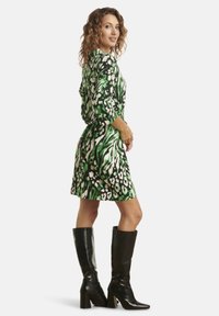 Grün und schwarz bedrucktes Kleid mit drei Viertelärmeln, Taillengürtel und knielangem Saum, kombiniert mit schwarzenkniehogen Stiefeln.