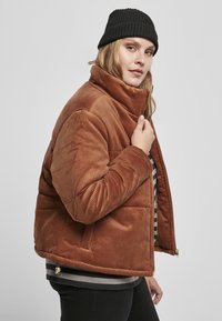 Bruine corduroy pufferjack met een hoge kraag en ritssluiting. Heeft een geribbelde textuur, zijzakken en een relaxed fit.