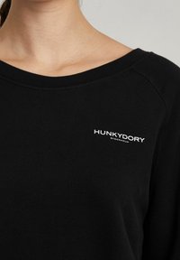 Schwarzer Sweatshirt mit weitem Halsausschnitt, aus weichem Material. Verziert mit dem weißen Logo "HUNKYDORY STOCKHOLM" auf der linken Brustseite.