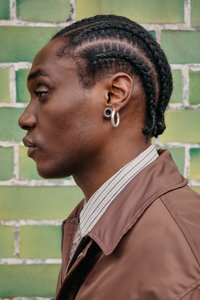 Profile view of a person wearing a brown zip jacket, a checkered shirt, and silver hoop earrings against a green and white brick wall.