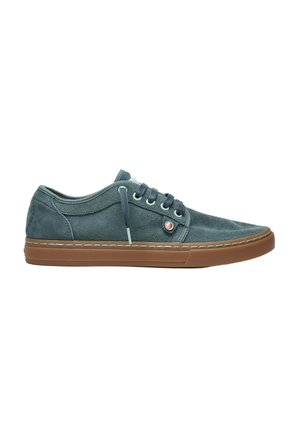 Zapatilla deportiva baja informal con parte superior de ante gris oscuro, cordones negros, ojales azul claro y suela de goma marrón sobre fondo blanco.