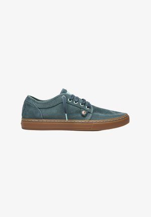Zapatilla deportiva baja informal con parte superior de ante gris oscuro, cordones negros, ojales azul claro y suela de goma marrón sobre fondo blanco.