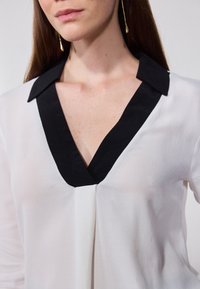Bílá blůza s černým límečkem, se širokým V výst�řihem a měkkou, jemně texturovanou tkaninou. Minimalistický design bez ozdob.