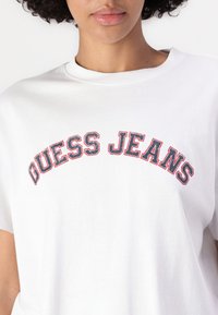 Baltas medvilninis marškinėliai su kreiva "GUESS JEANS" logotipu raudona ir mėlyna spalvomis. Apvalaus kaklo dizainas su trumpomis rankovėmis, glotnus paviršius.