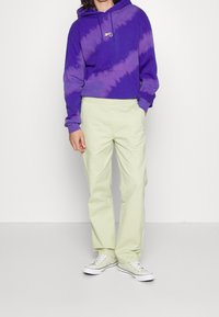 Violetin kirjava huppari, jossa on etulogo, yhdistettynä vaaleanvihreisiin housuihin. Mallilla on päällään valkoiset tennarit, joissa on kuviollinen design.