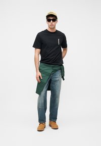 Νεαρός άνδρας που φοράει μαύρο μπλουζάκι Timberland, πράσινο φούτερ δεμένο στη μέση, ξεθωριασμένο τζιν, ταμπά μπότες, γυαλιά ηλίου και κίτρινο καπέλο με φόντο το λευκό.